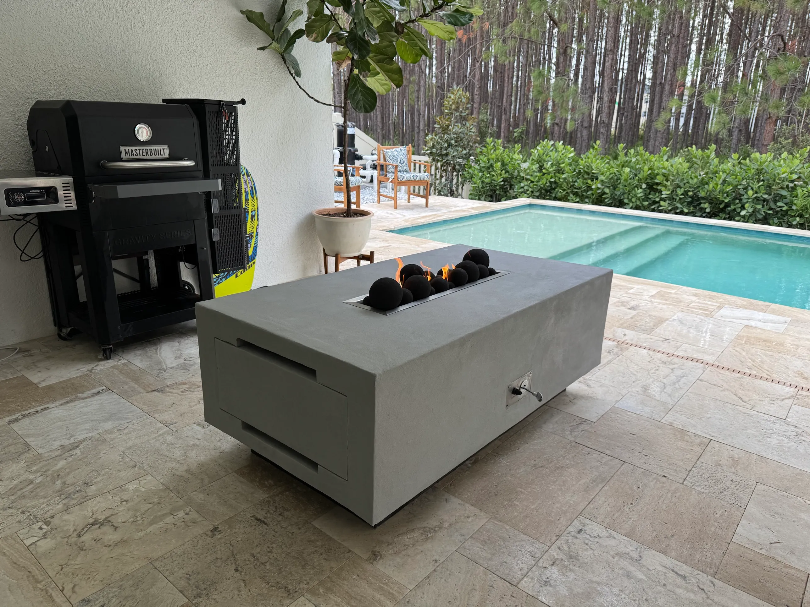 A concrete-grey fire table with black fire stones, on a stone patio beside a pool